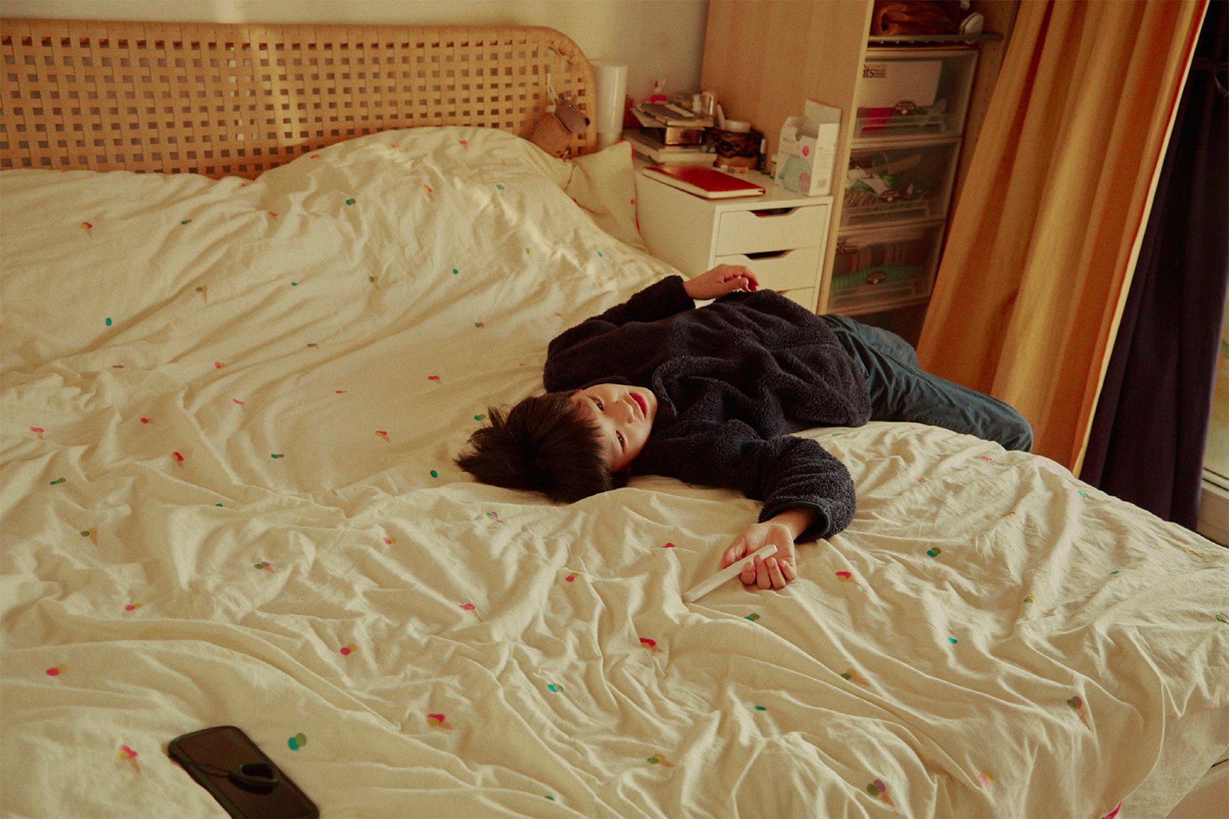 Young adult lies on a patterned bed in an indoor room, holding a white object. Phone and bedside table nearby.
