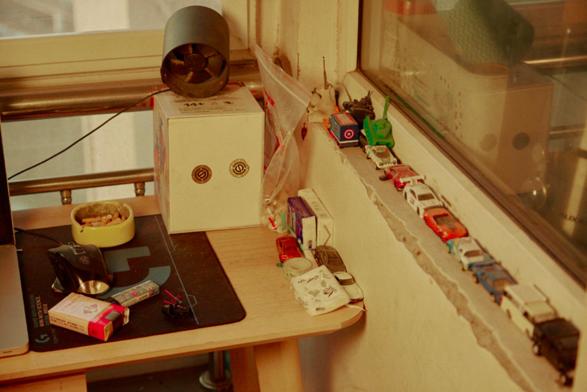 Still life of objects on a desk including a vintage fan, toy cars, and cigarettes in front of a window, creating a nostalgic scene.
