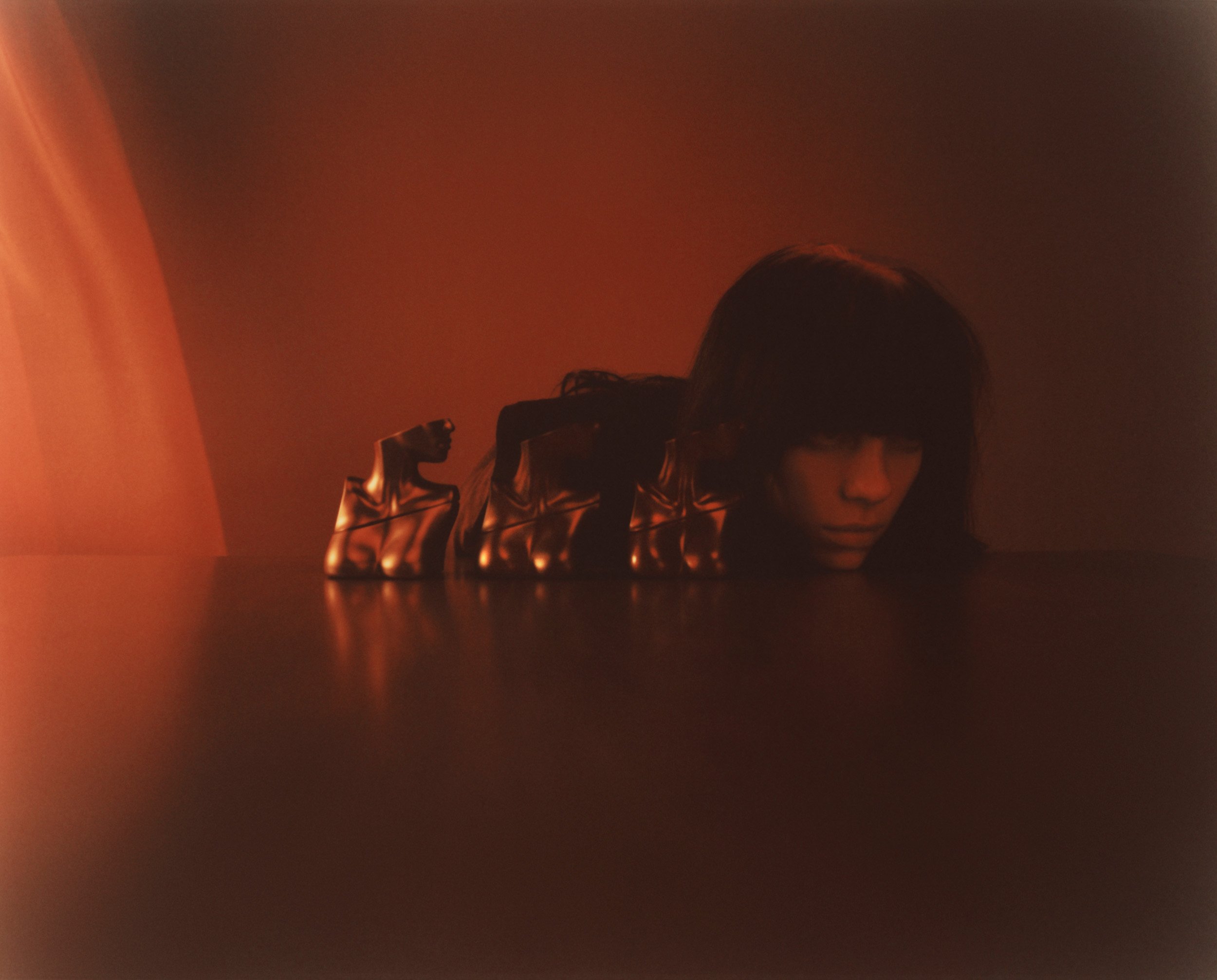 Monochromatic portrait of a woman with blunt bangs behind a table, wearing gold gloves with molded faces on the fingers in soft lighting.