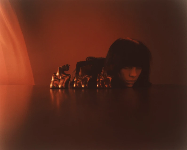 Monochromatic portrait of a woman with blunt bangs behind a table, wearing gold gloves with molded faces on the fingers in soft lighting.