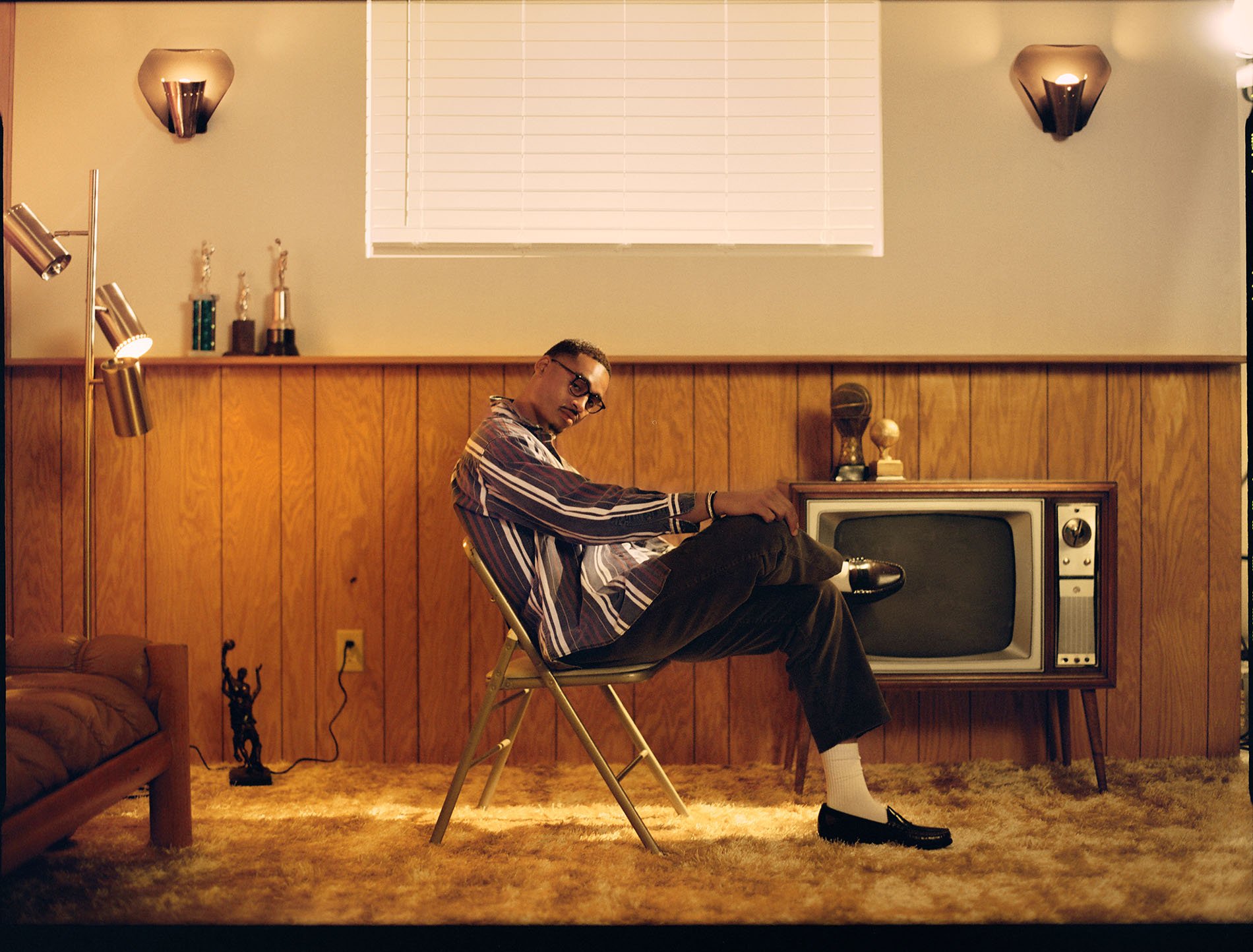 Adult male sitting on a folding chair in an indoor living room setting with a vintage television, lamp, trophies and wood paneled walls.