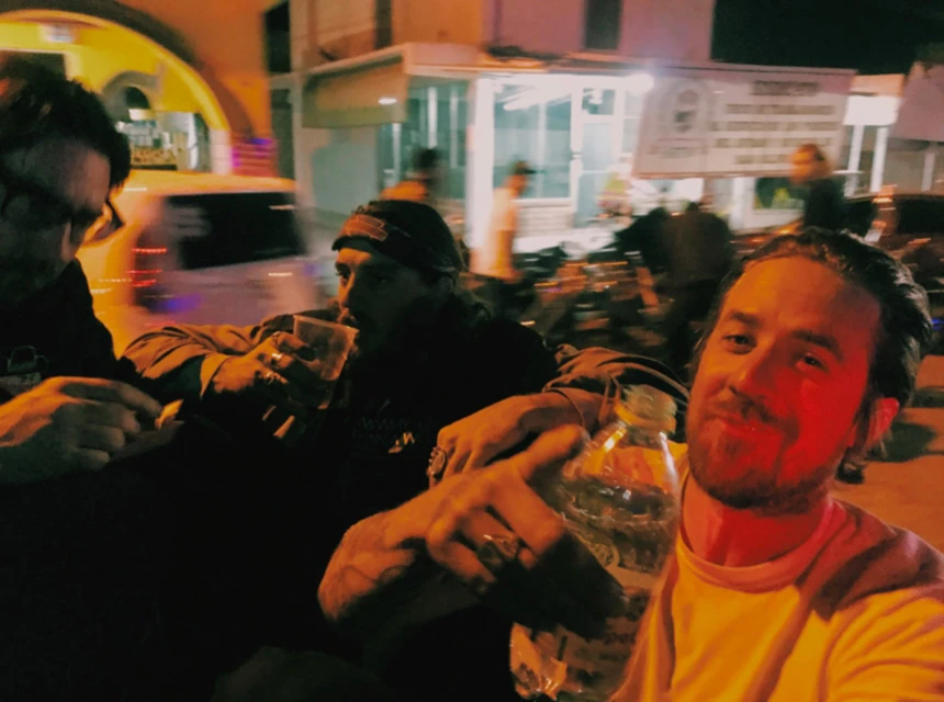 Three adult males socializing outdoors at night, drinking from cups and a water bottle in an urban area with buildings in the background.