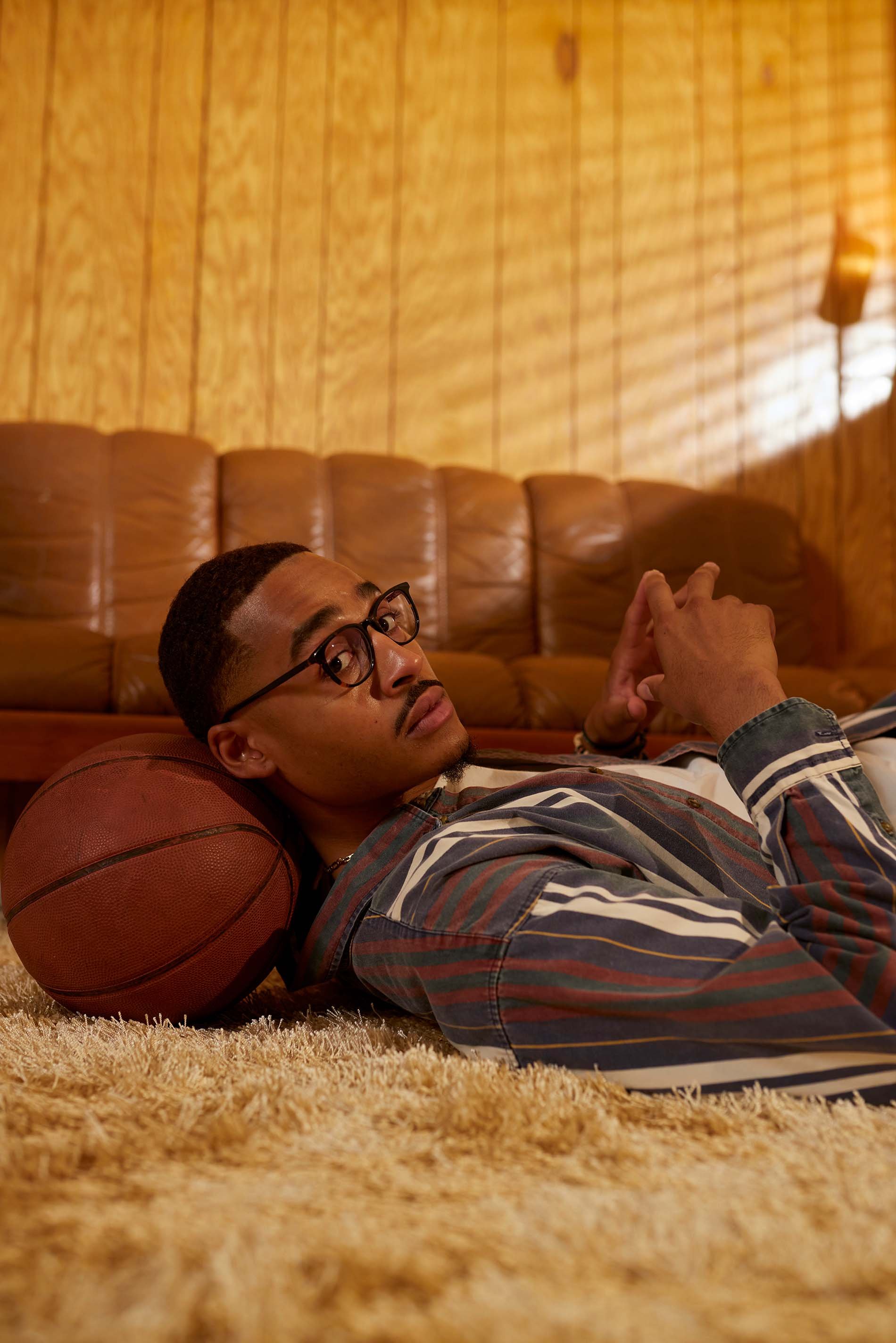 An adult male lies on a shag carpet next to a basketball in a retro interior setting with wood paneling and a tan leather sofa.