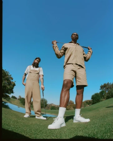 Two men in matching tan outfits and white shoes pose on a sunny golf course with one holding a golf club across his shoulders.