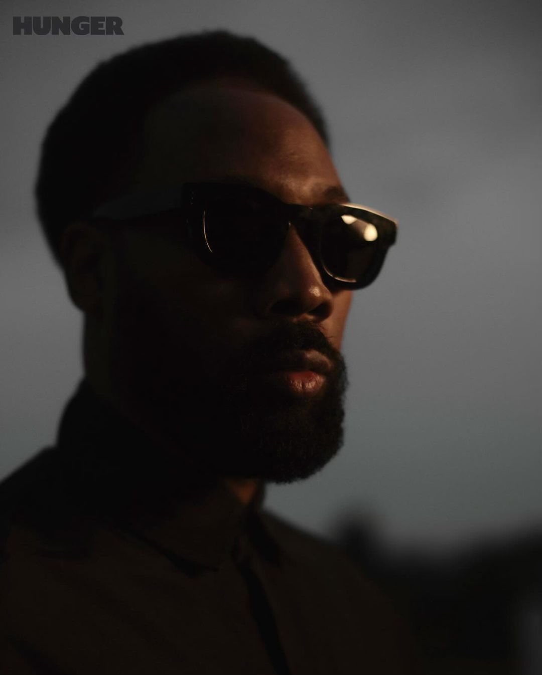 Close-up portrait of an adult male wearing sunglasses, with a beard, against a blurred gray background, with 'HUNGER' logo in corner.
