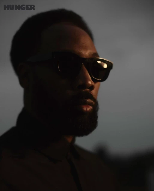 Close-up portrait of an adult male wearing sunglasses, with a beard, against a blurred gray background, with 'HUNGER' logo in corner.