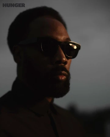 Close-up portrait of an adult male wearing sunglasses, with a beard, against a blurred gray background, with 'HUNGER' logo in corner.
