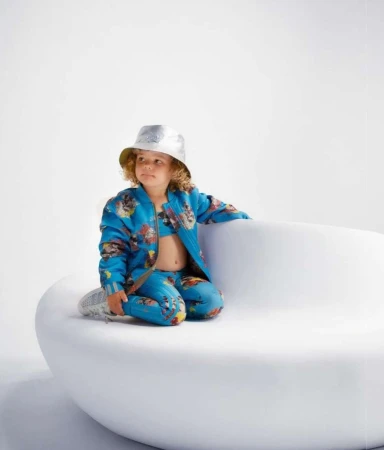 Child model wearing a blue patterned tracksuit and silver bucket hat seated on a white curved sofa
