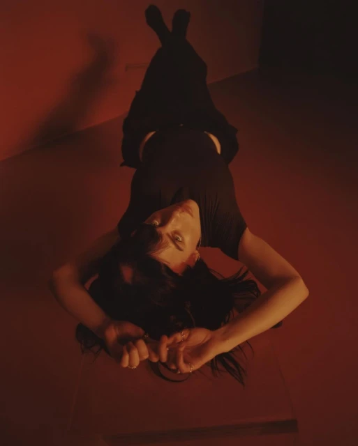 Dark-haired young adult woman in a black top and skirt lies on her back in a red room, gazing upwards with a slightly smiling expression.