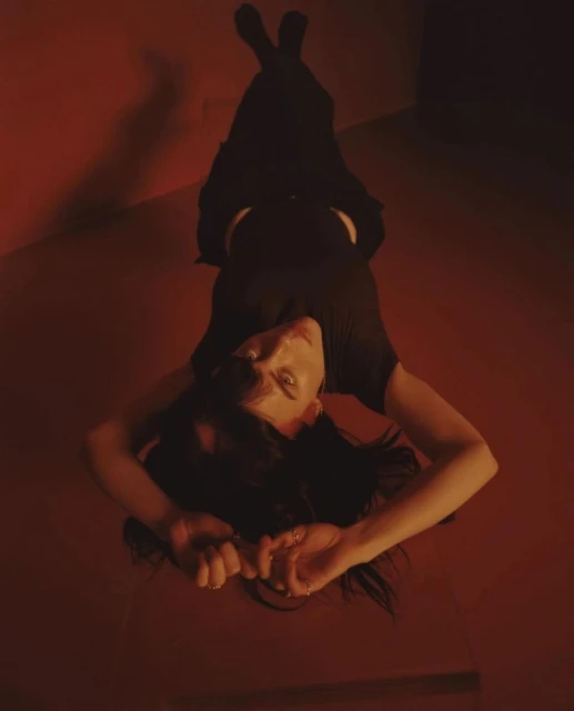 A woman in a dark outfit is lying on a red floor in a corner looking up at the camera in a dramatic, high-angle, indoor portrait.