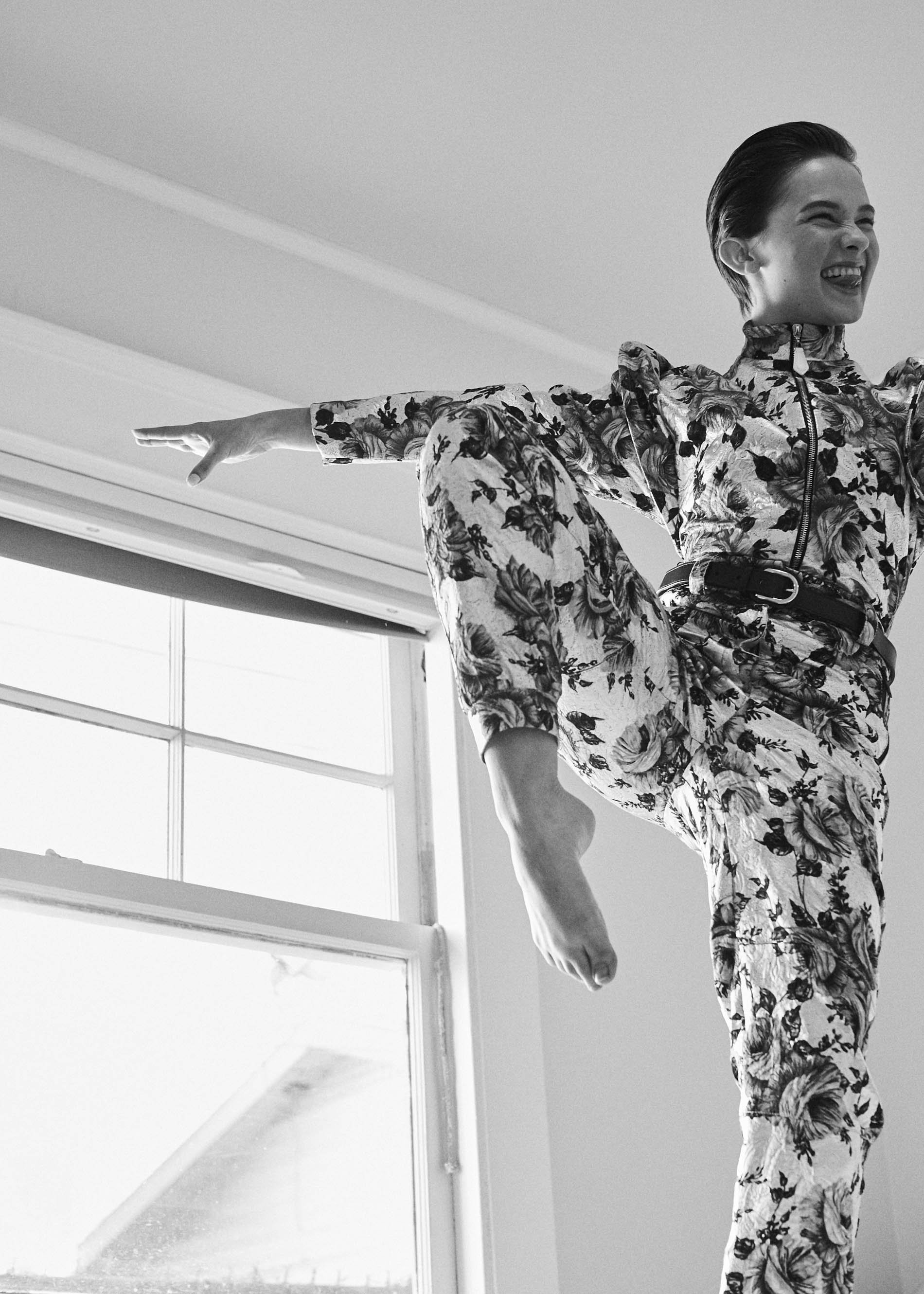 A person in a floral jumpsuit strikes a pose mid-air near a window in a monochrome, indoor setting, showcasing fashion and lifestyle.