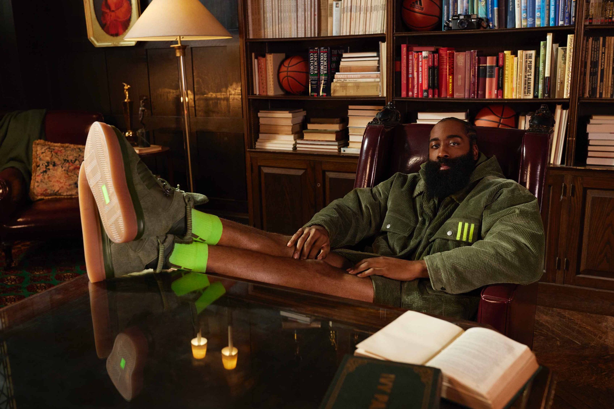 An adult male with a beard sits in a leather chair, legs up on a table in a corduroy jacket and bright green socks in a room with a bookshelf.
