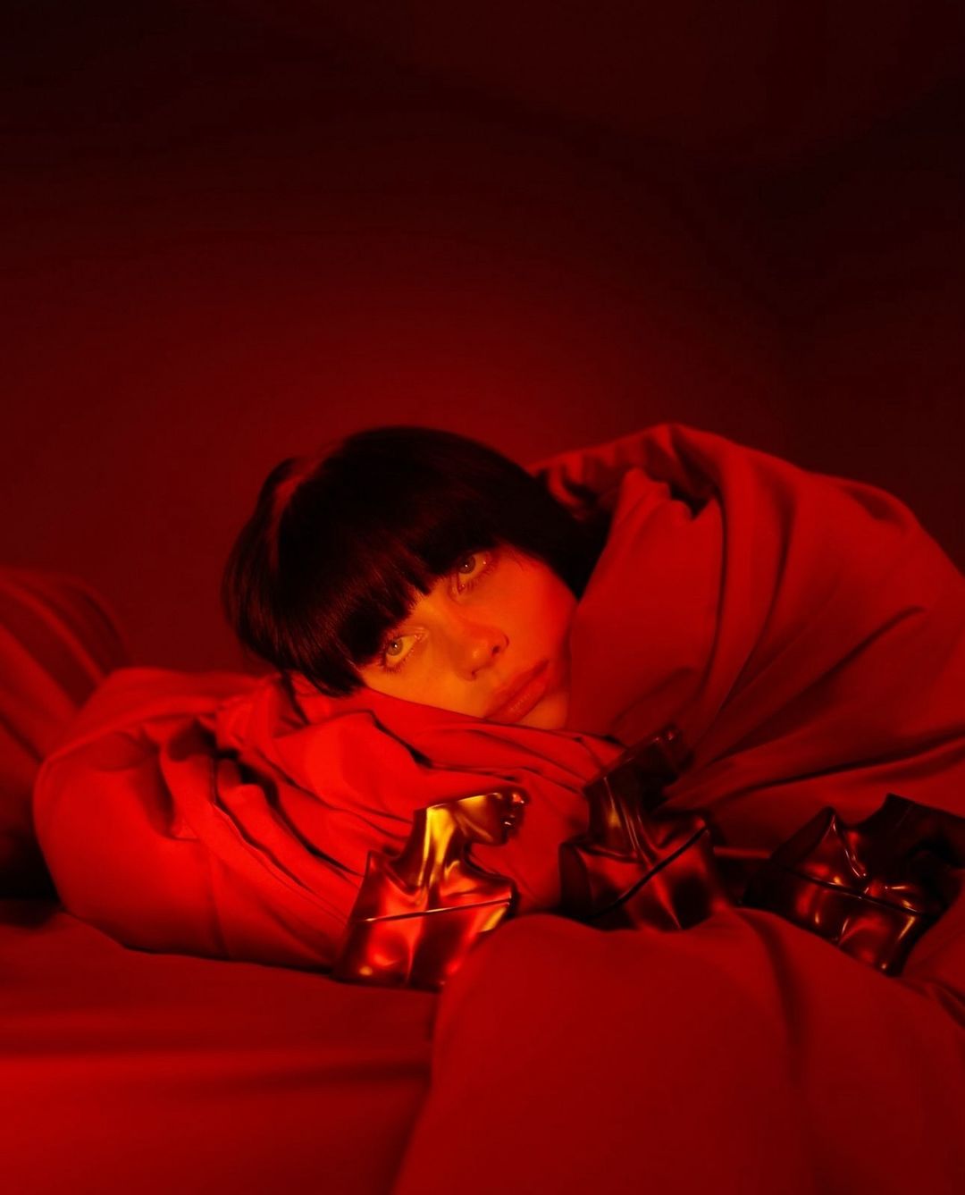 Adult female with dark hair, lying down on a red surface, partially covered by red fabric, with small golden objects in the foreground.