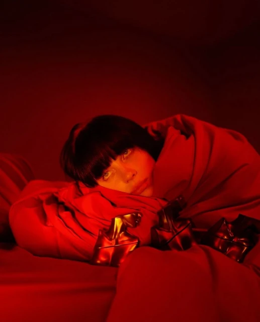 Adult female with dark hair, lying down on a red surface, partially covered by red fabric, with small golden objects in the foreground.