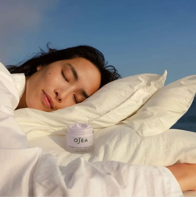 Woman sleeping on a pillow with a jar of Osea collagen night cream on the bedding, set against a blue sky backdrop.
