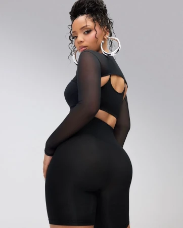 Full body studio shot of an adult female wearing a black workout outfit with a mesh top and large hoop earrings against a grey background.