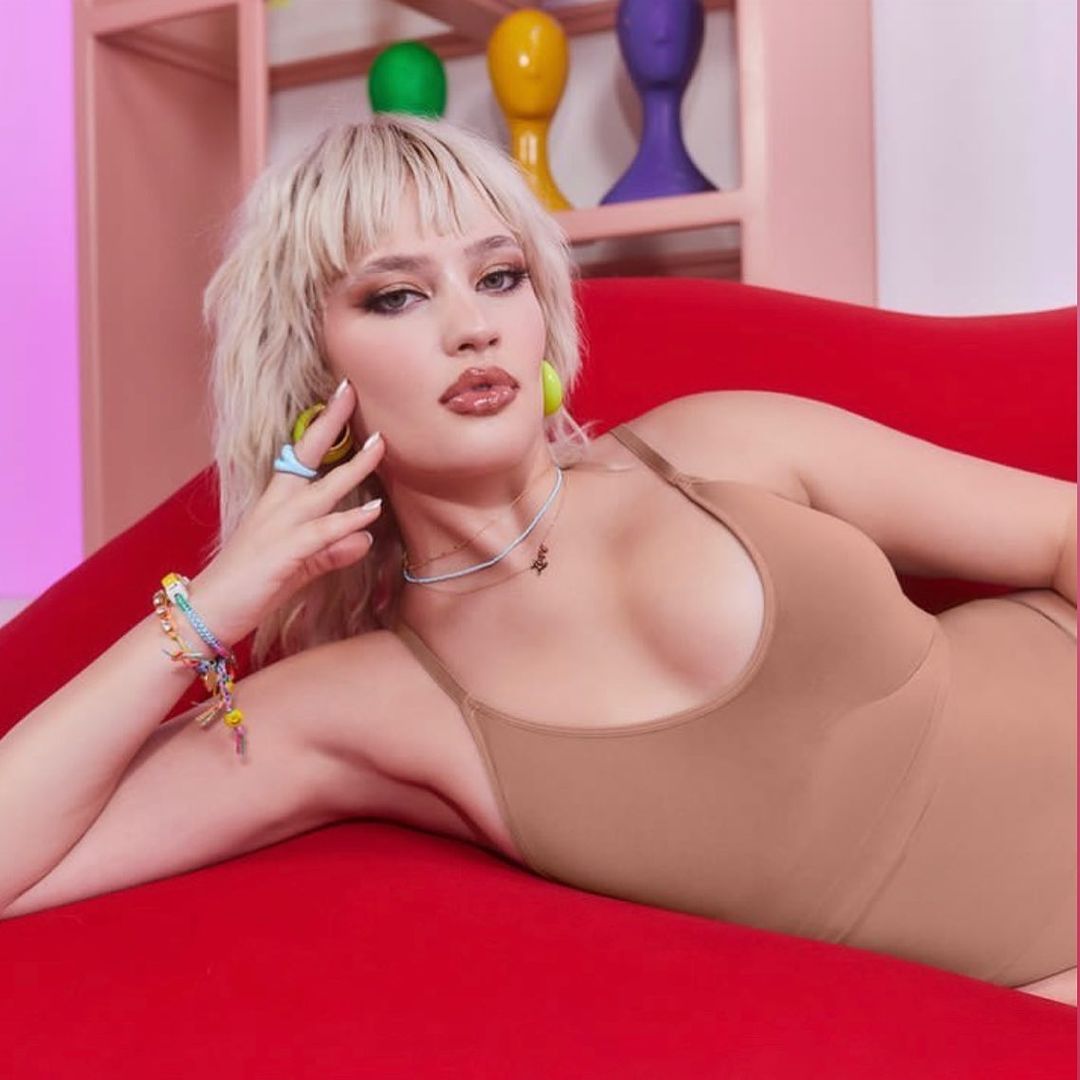 Blonde woman reclines on a red couch, gazing at the camera in a close-up shot. She wears a beige bodysuit and colorful jewelry.