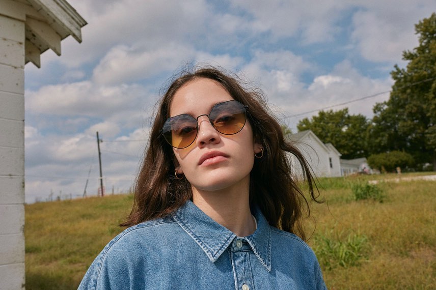Woman wearing gradient sunglasses and a blue denim shirt outdoors against a rural sky