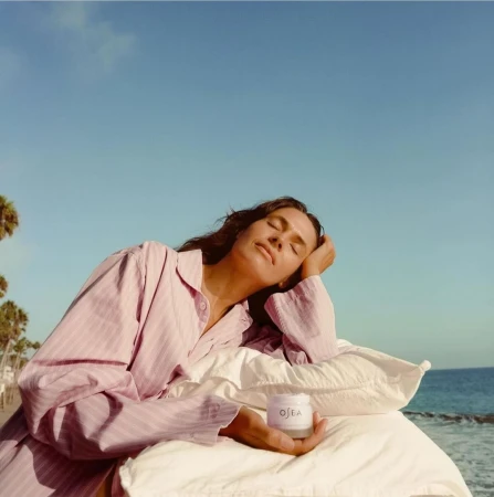 Woman in striped pink shirt resting on white pillows holding a jar against an ocean and clear sky background