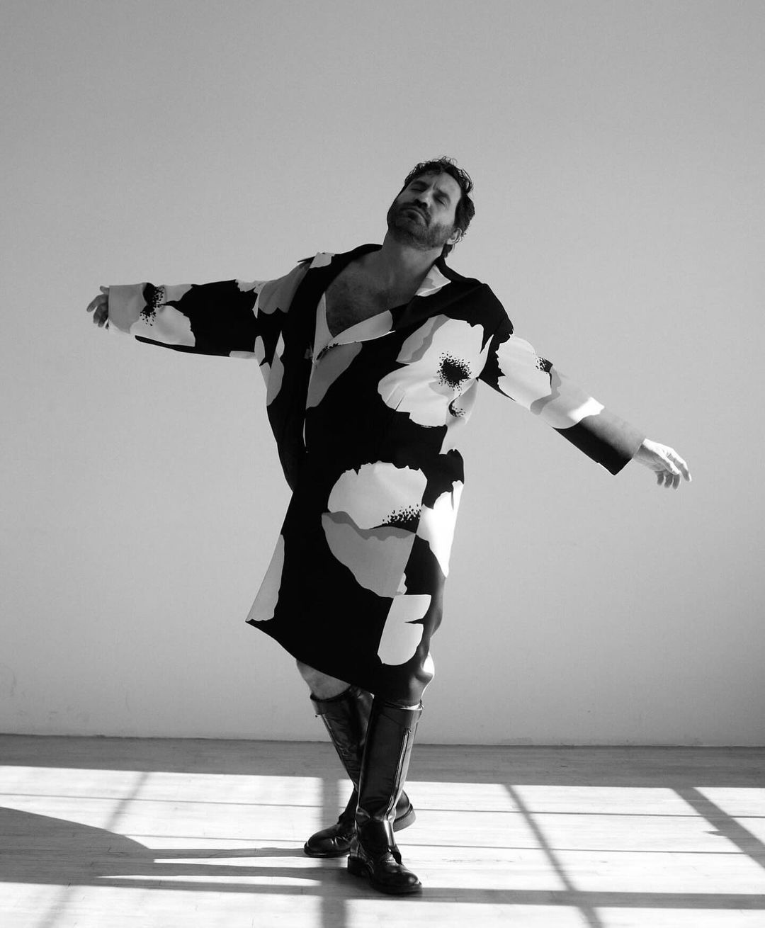 Black and white studio shot of a man in a floral print coat and knee-high boots, arms outstretched, eyes closed, standing on a wood floor.