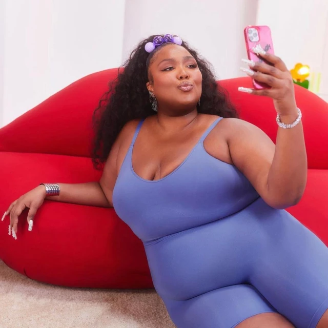 Lizzo poses with a pink smartphone against a backdrop featuring a large red lip couch and white walls in a vibrant indoor setting.