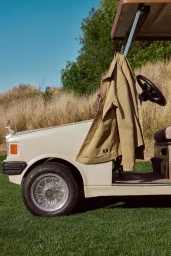Cream golf cart parked on green turf with a beige jacket hanging from the roof support under a clear blue sky