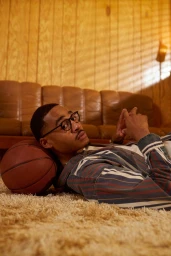 A man wearing glasses and a striped shirt lying on a carpet with his head resting on a basketball.