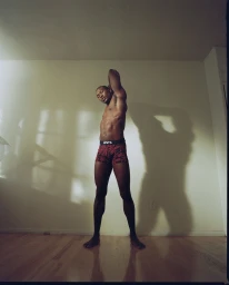 Black man stretching in patterned underwear with arms raised inside a room with light wooden floors.