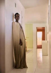 Black woman wearing a long gold hooded gown leaning against a white hallway wall in a Los Angeles interior by photographer Robin Harper