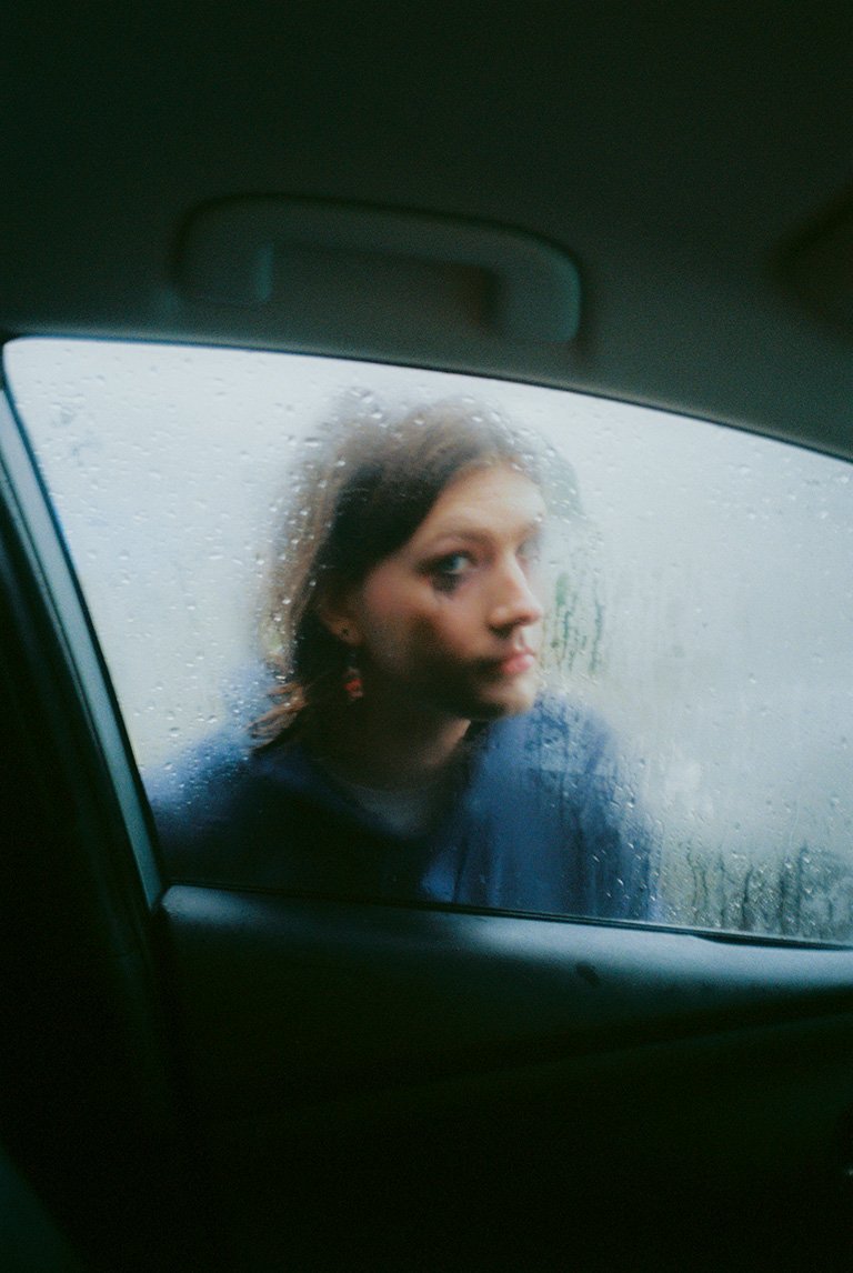 Woman with dark hair sits in car on rainy day, gazing through the window with raindrops. Vehicle interior and rear window visible.