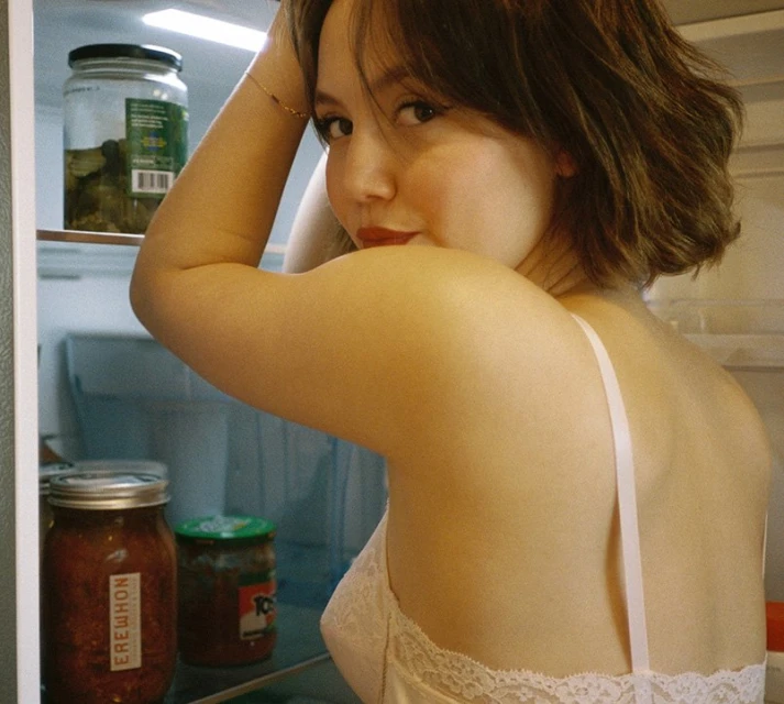 Woman wearing a white lacy top looks over her shoulder in an indoor environment; the background includes glass jars in a refrigerator.