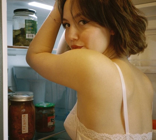 Woman wearing a white lacy top looks over her shoulder in an indoor environment; the background includes glass jars in a refrigerator.