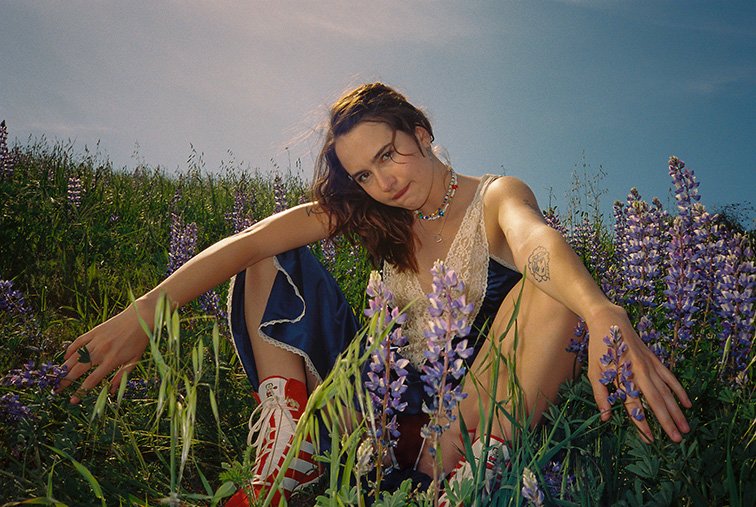 Young woman in a navy dress with lace trim sitting in a field of purple lupine flowers, wearing red and white sneakers and a beaded necklace.