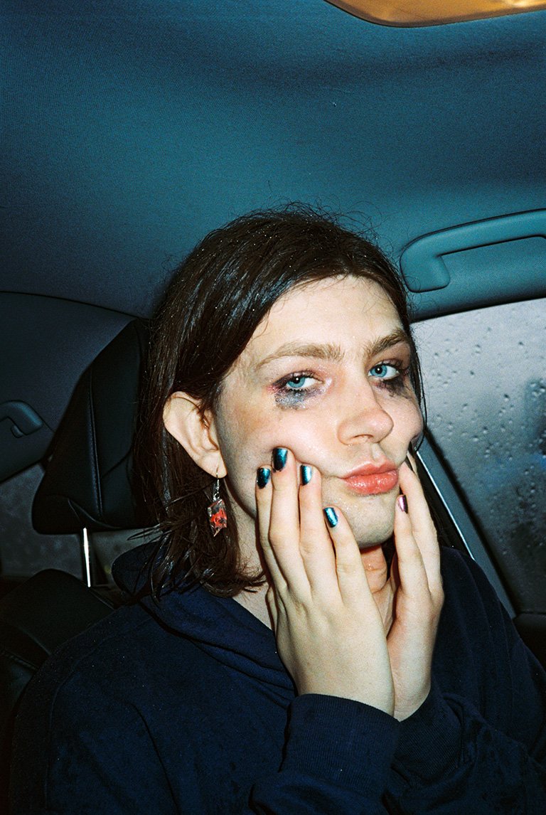 Close-up of a person in a car with smeared makeup, touching their face, and wearing a dark hooded top, capturing a raw and emotional portrait.