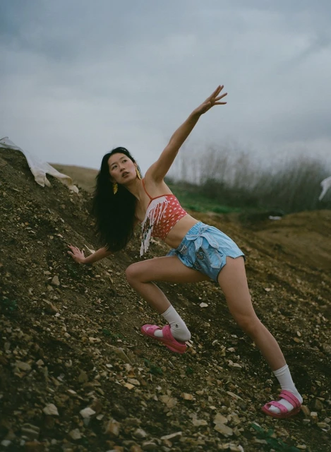 Young Asian woman in unique fashion attire climbs a dirt hill on a cloudy day, showcasing a blend of style and natural setting.