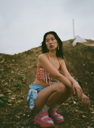 East Asian woman in red star-print crop top and blue shorts squatting outdoors on a dirt mound under an overcast sky.