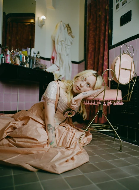 Woman with blonde hair reclines in pink dress, leaning against a pink vanity chair in a pink-tiled bathroom with cosmetics displayed on the counter.