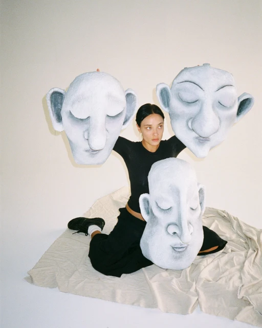 Woman sits among three large paper mache heads, each with closed eyes, neutral expressions, and prominent ears, indoors in soft light.
