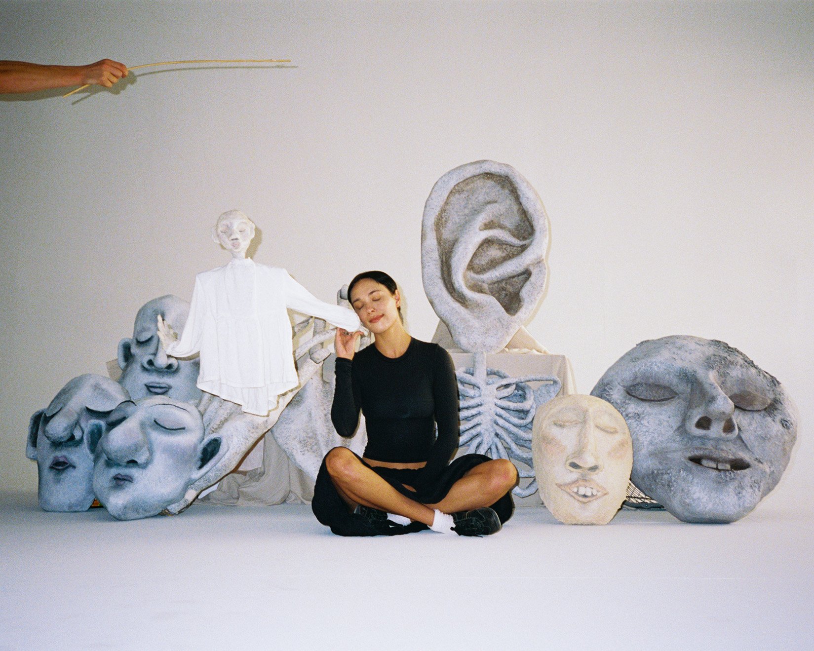 Woman seated amid artful head sculptures and a skeleton prop in a bright studio setting, with a pair of hands and sticks above.