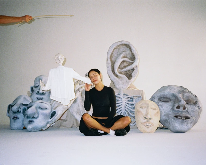 Woman seated amid artful head sculptures and a skeleton prop in a bright studio setting, with a pair of hands and sticks above.