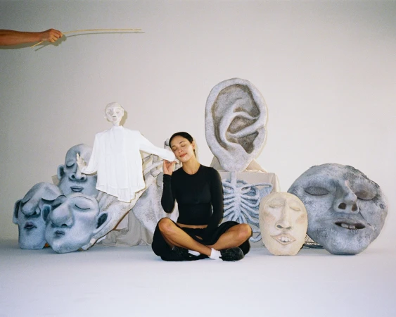 Woman seated amid artful head sculptures and a skeleton prop in a bright studio setting, with a pair of hands and sticks above.