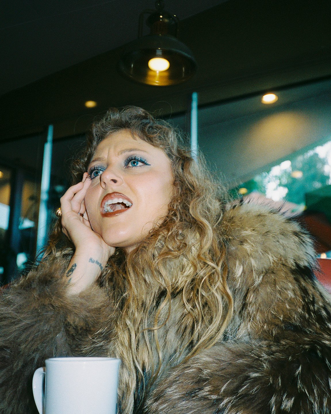Woman in a fur coat sits indoors with a coffee cup, looking upwards toward the light fixture with an intrigued facial expression.
