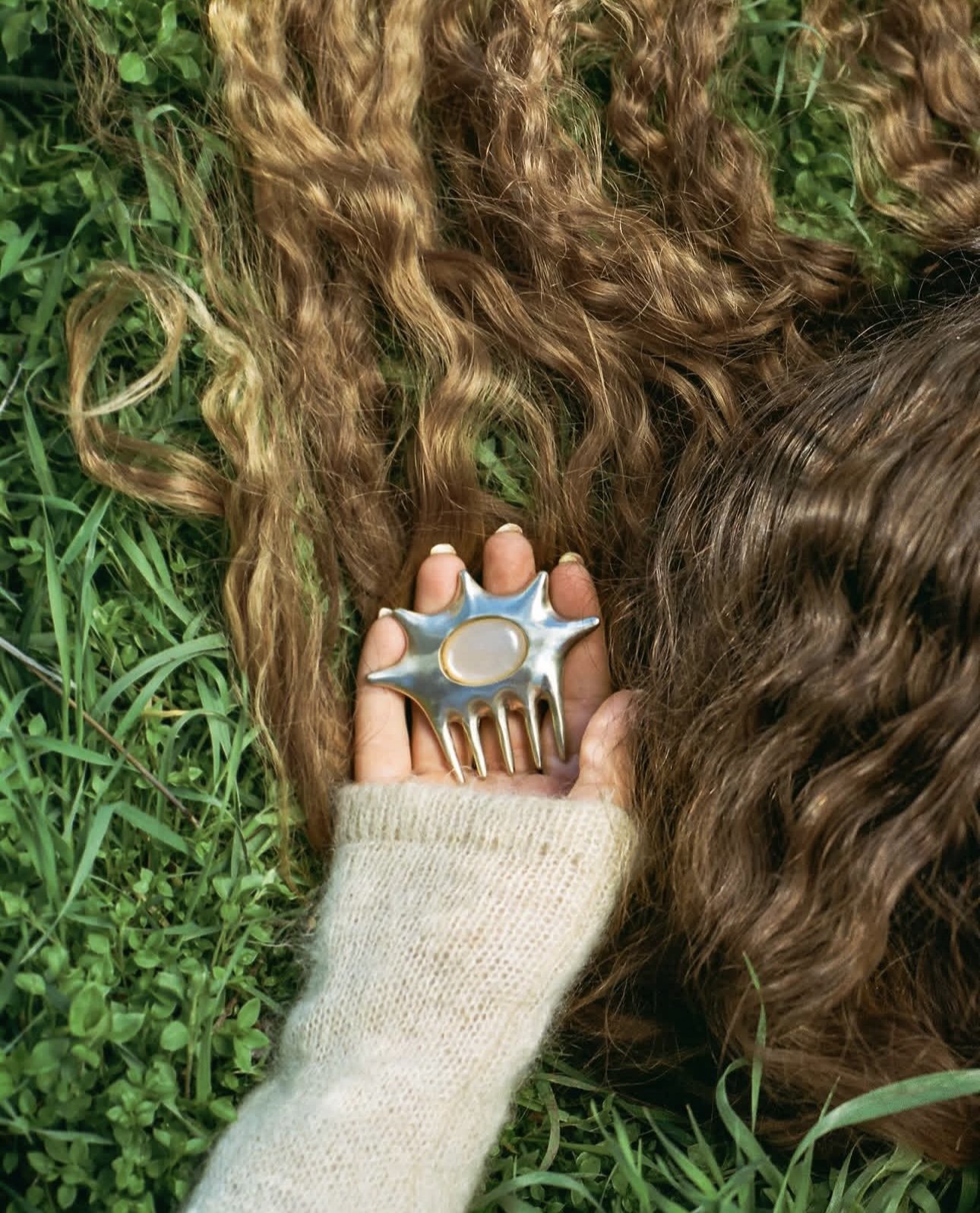 Woman's hand holds a star-shaped silver comb with an opal stone while lying on grass, showcasing fashion and nature.
