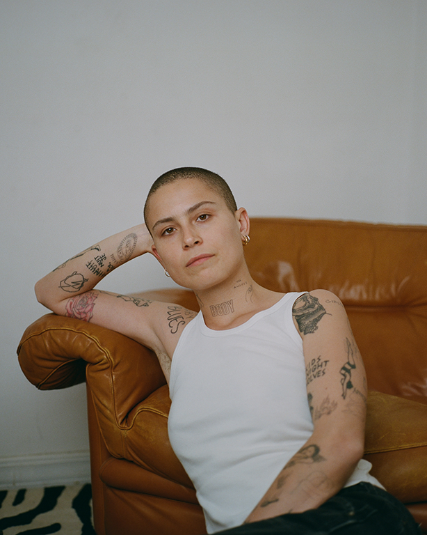 Portrait of a young adult with tattoos in a white tank top sitting on the floor next to a tan leather couch in an indoor environment.