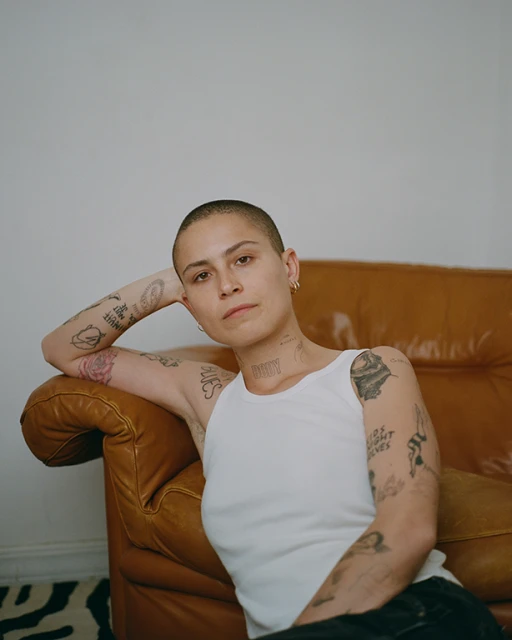 Portrait of a young adult with tattoos in a white tank top sitting on the floor next to a tan leather couch in an indoor environment.