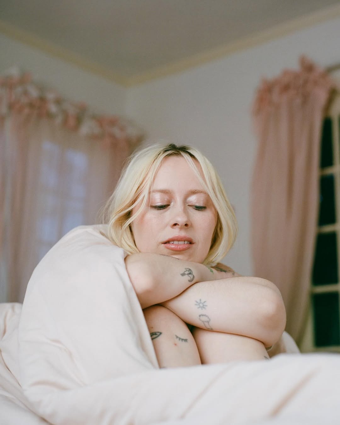 Woman with blonde hair and arm tattoos sits wrapped in a blanket indoors near window with light pink curtains and natural lighting.