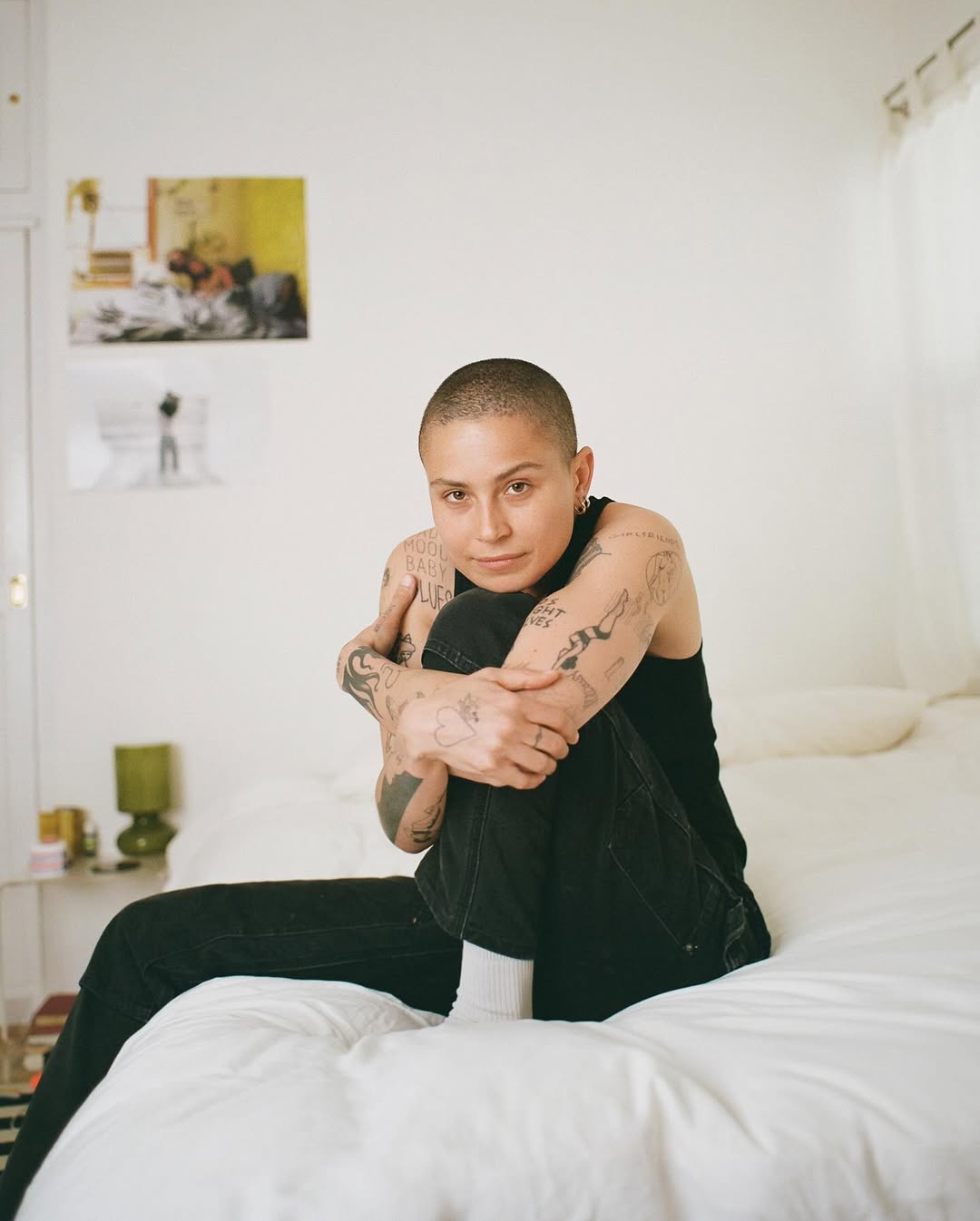 Adult woman with tattoos sits cross-legged on a bed in a bright indoor bedroom with white walls and natural lighting from a window.