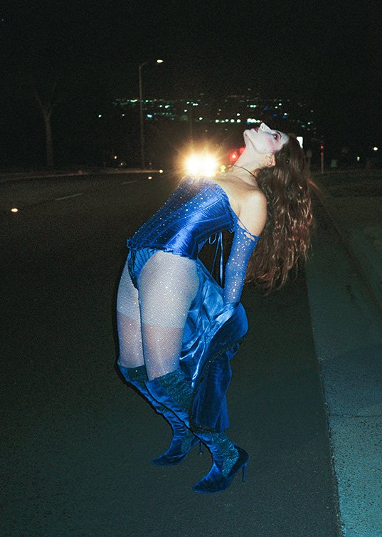 Woman in blue sequined bodysuit and thigh-high boots poses on a dark road at night, illuminated by car headlights and city lights.