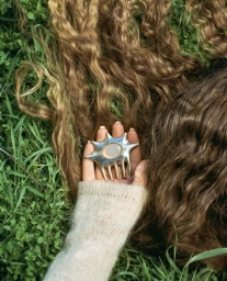Close-up of a hand holding a metallic star-shaped comb resting on grass near long brown hair, photographed by Zoe Donahoe.