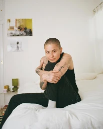 Portrait of a tattooed person with a buzzcut seated on a white bed wearing a black tank top and dark pants, photographed by Zoe Donahoe.